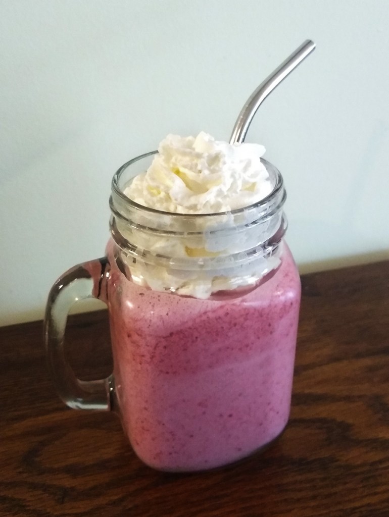 A photo of a mason jar style mug with a pink smoothie in it, topped with whipped cream and a metal straw.